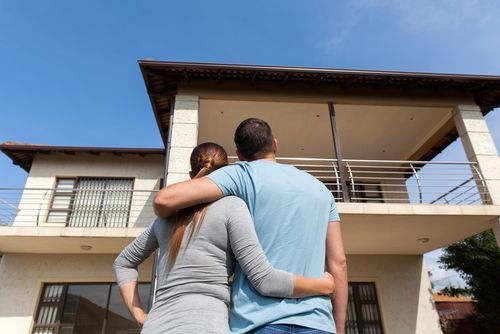 A man and woman stand together in front of a house, smiling and looking towards the camera.