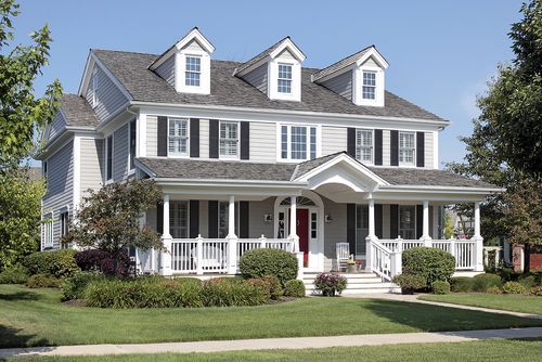 Colonial-style house with three dormer windows. The porch has railings and is surrounded by lush greenery.