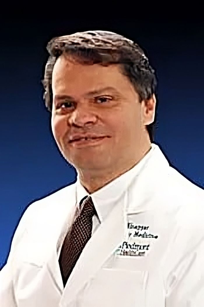 A man in a white coat and tie is standing in front of a blue background.