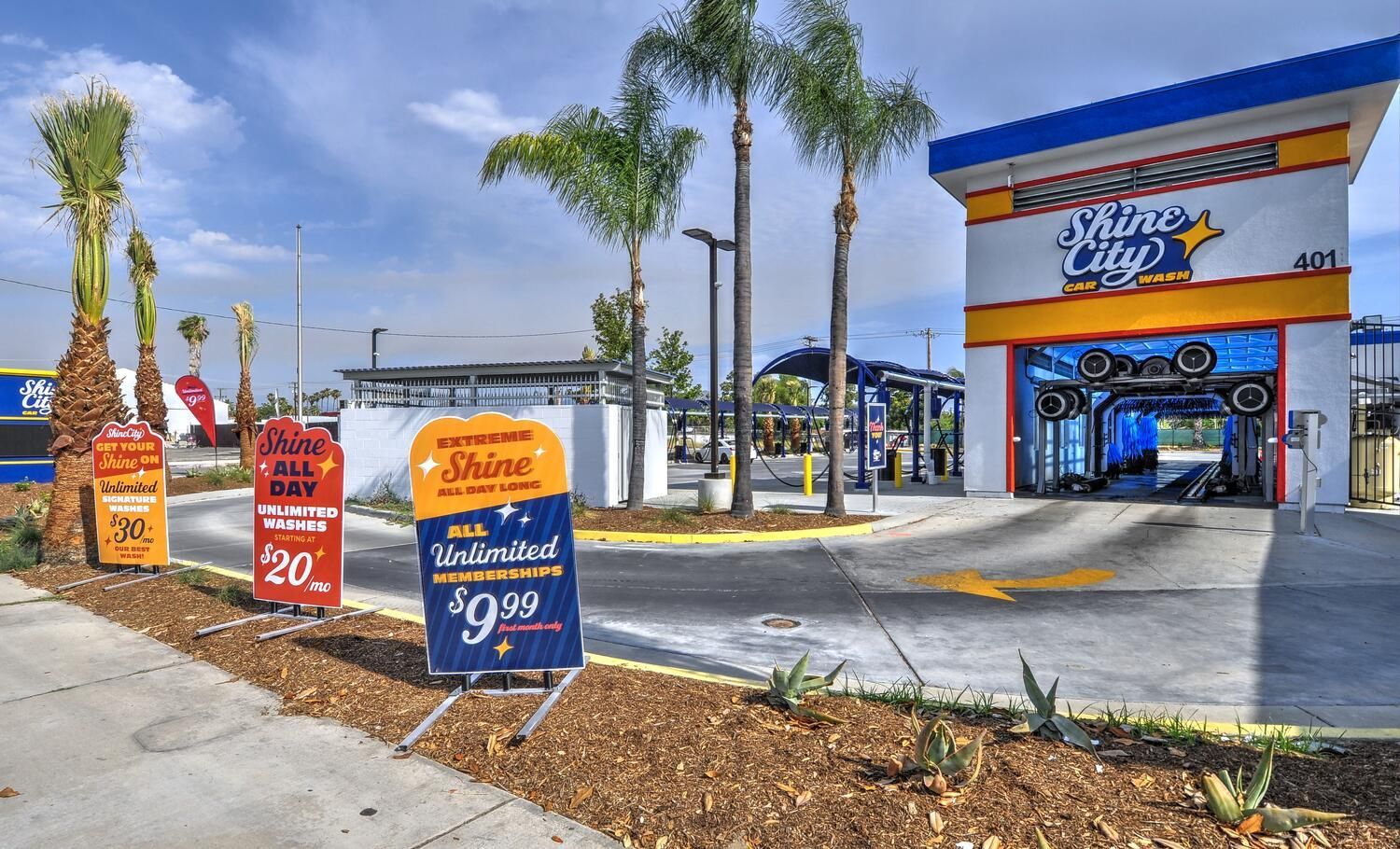 Exterior of a car wash facility; signs display wash packages. Palm trees and blue sky backdrop.