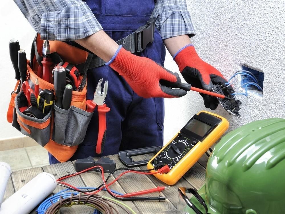 Electrician in blue overalls with red gloves, using a screwdriver on an outlet. Tools and equipment visible.