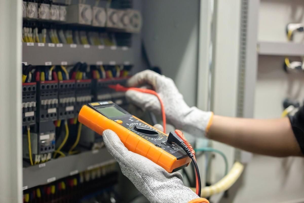 An electrician in safety gloves using a multimeter to inspect an electrical panel.