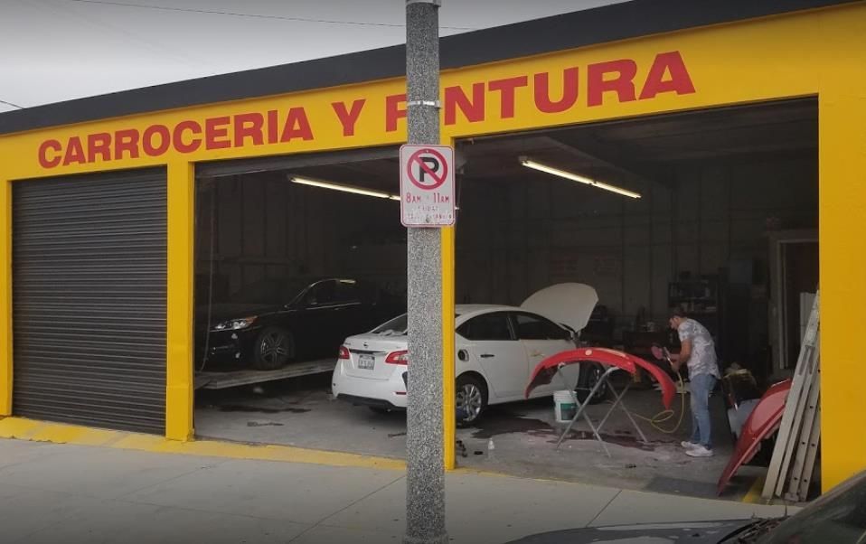 Yellow auto body shop with open bay doors. A person works on a red bumper. Cars inside.