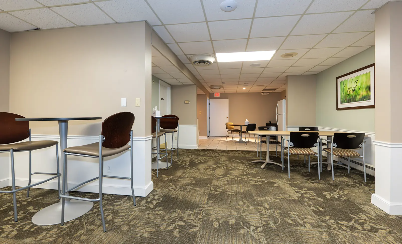 Communal lounge with tables, chairs, and carpet. Light brown and beige walls, with a doorway to another room.