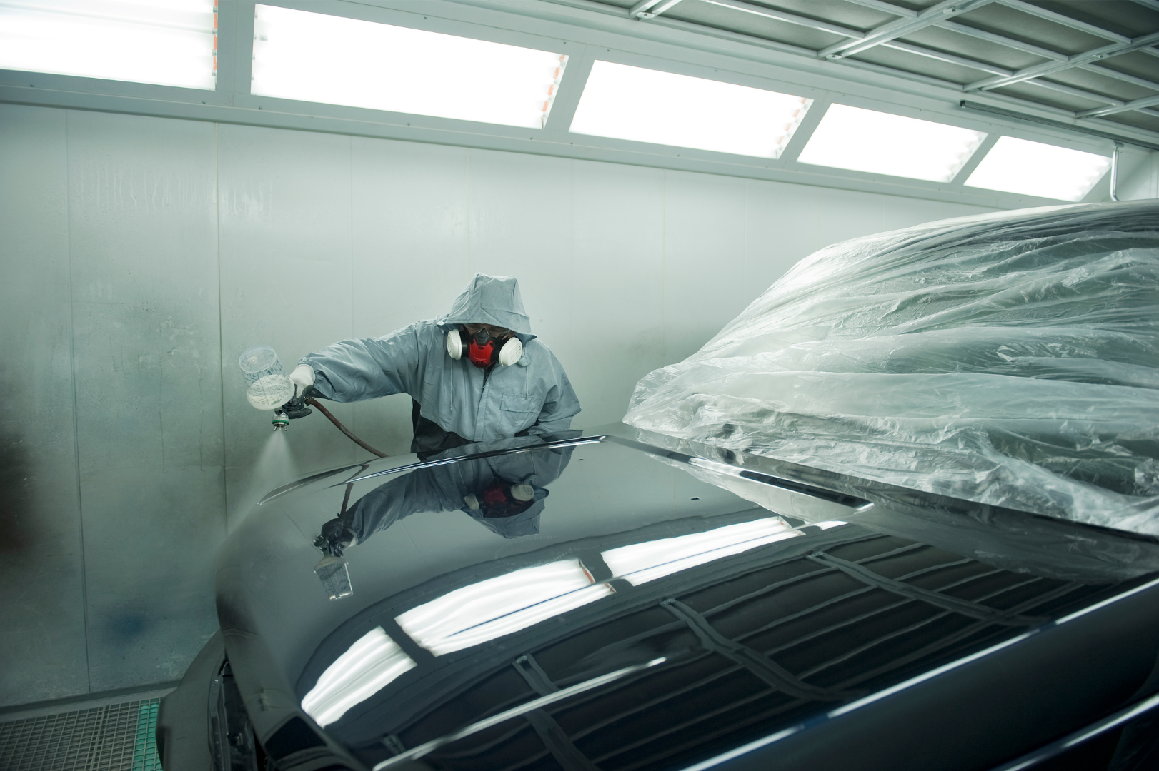 Worker in protective gear spray-painting a dark car in an auto body paint booth