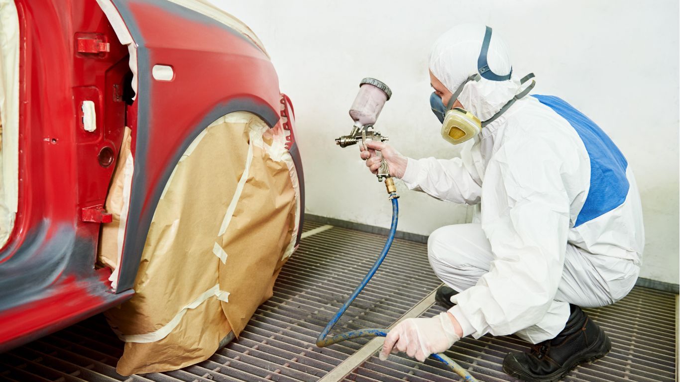 Worker in white protective suit spray-painting a red car panel in a paint booth