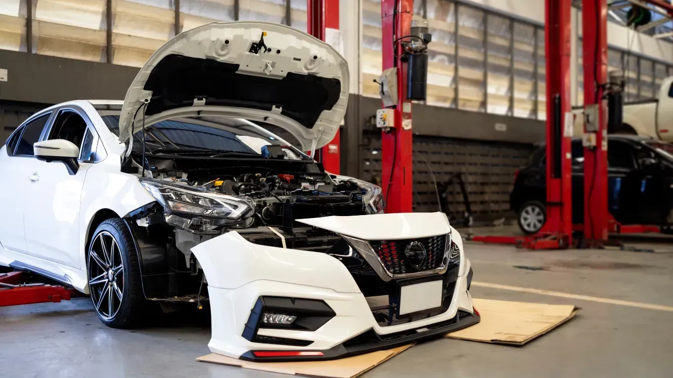 White car with hood open in an auto shop after front-end damage, near a red lift and tools.
