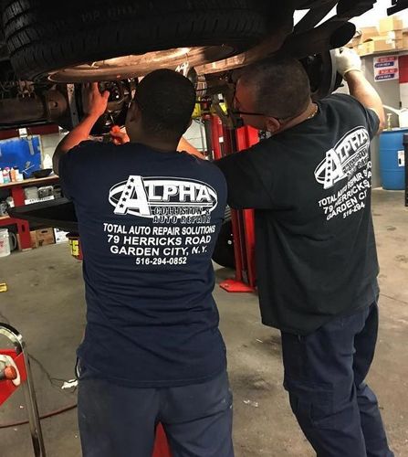 Two mechanics working under a raised car in a garage, wearing dark shirts with “ALPHA” on the back.