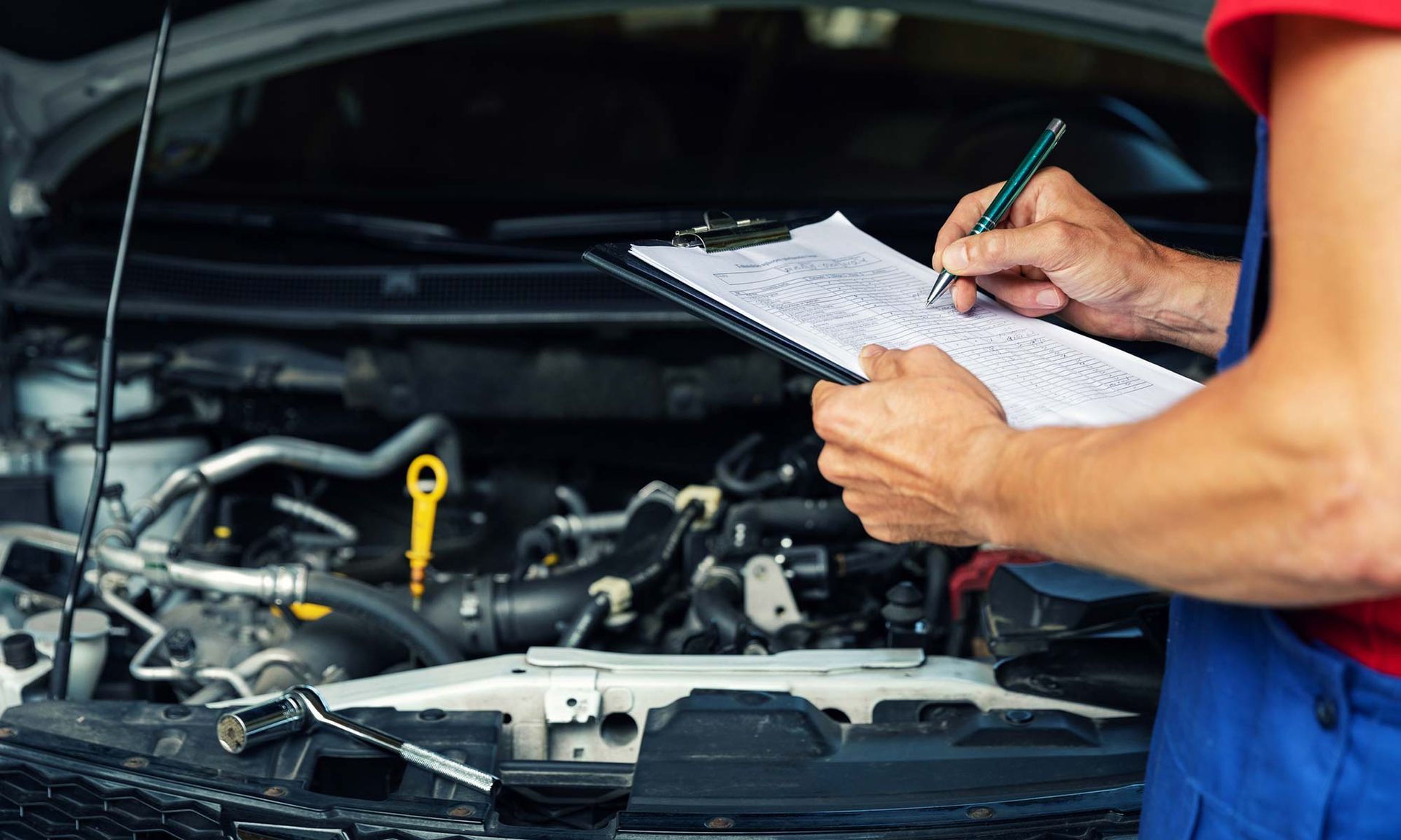 Mechanic writing on a clipboard beside a car engine with the hood open