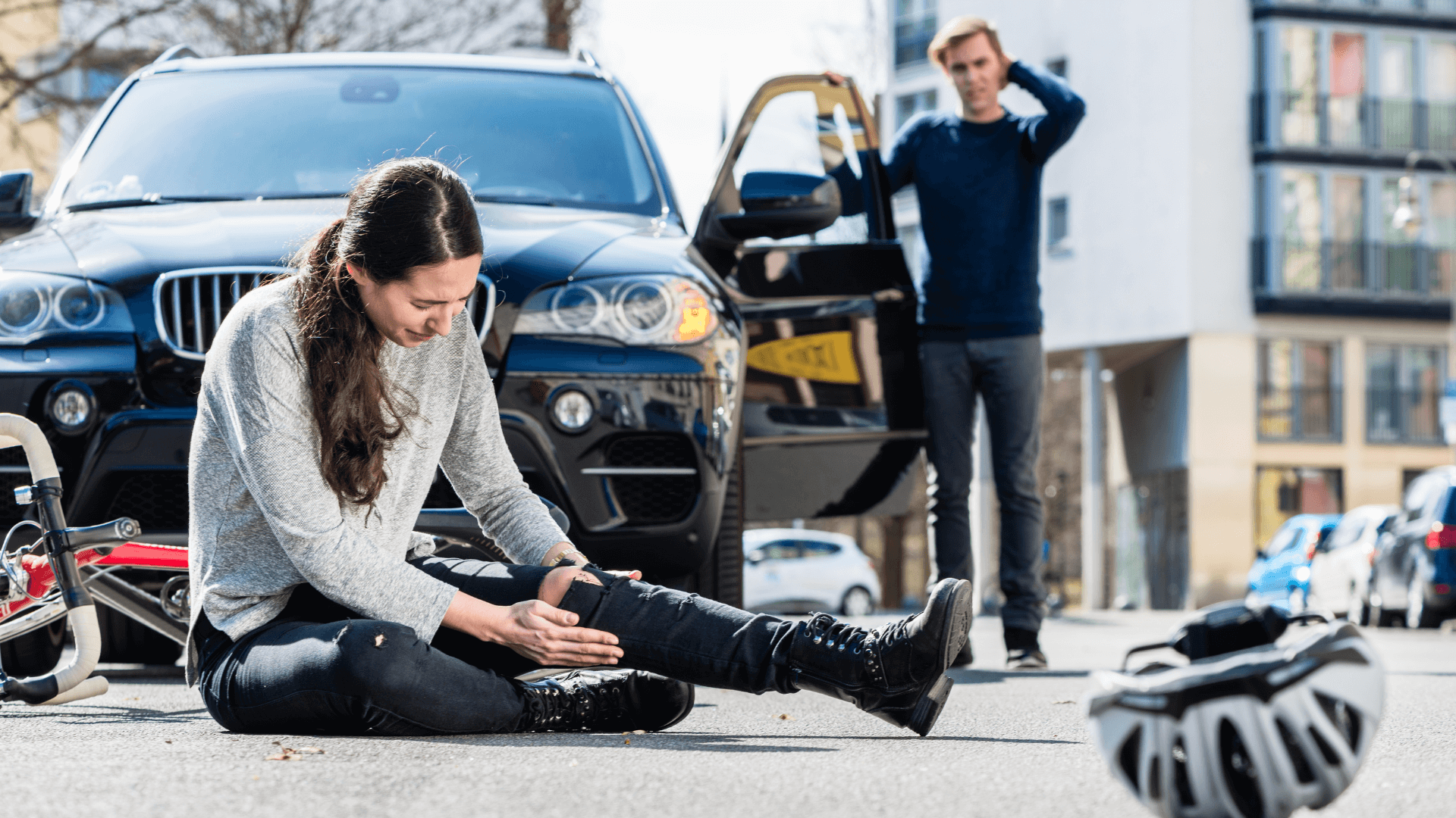 Person sitting on road, clutching an injured leg, with a car and worried bystander nearby.