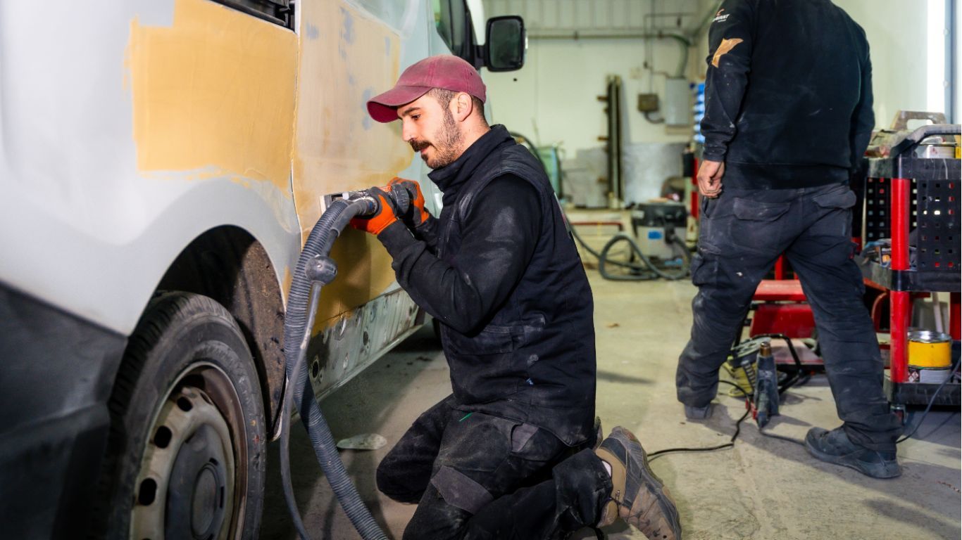 Two mechanics repairing a white van inside a garage, one kneeling with a power tool.