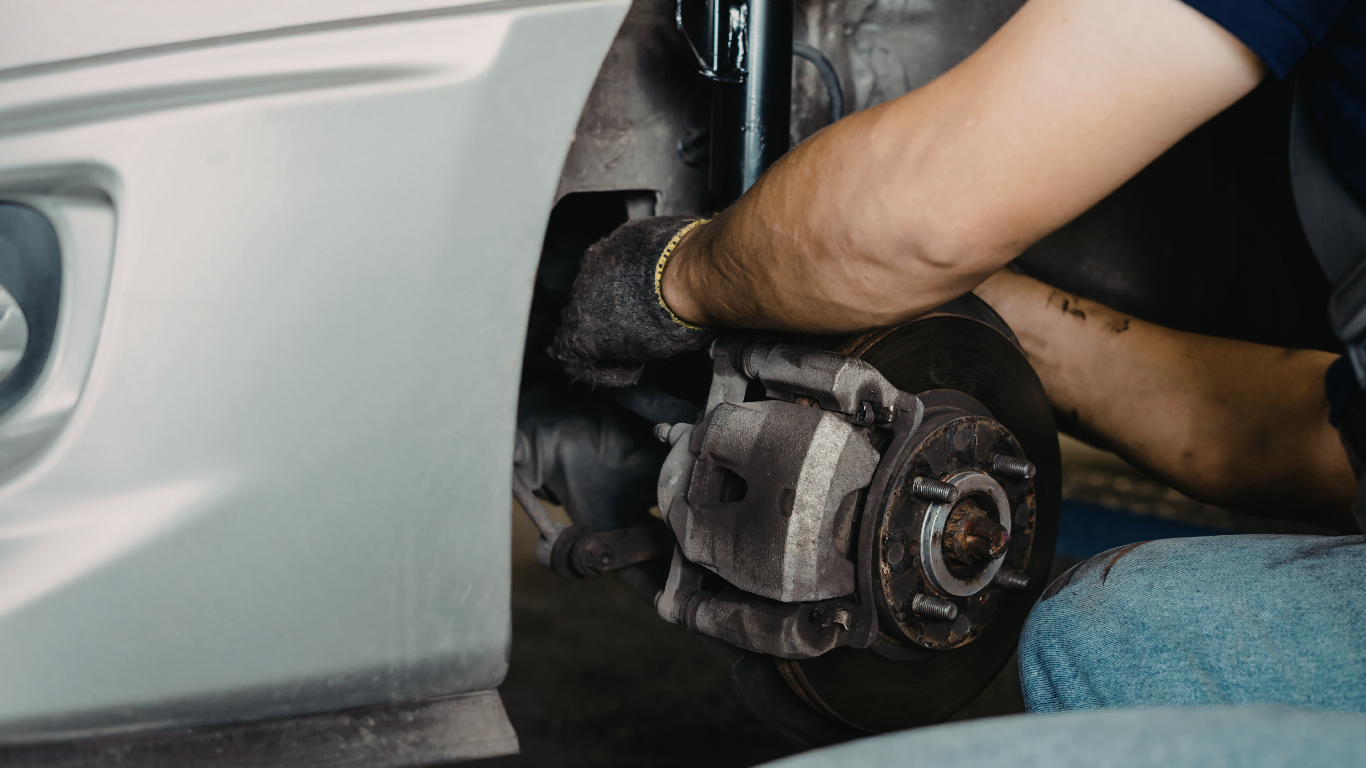 Mechanic replacing a car’s brake rotor and caliper near a silver vehicle