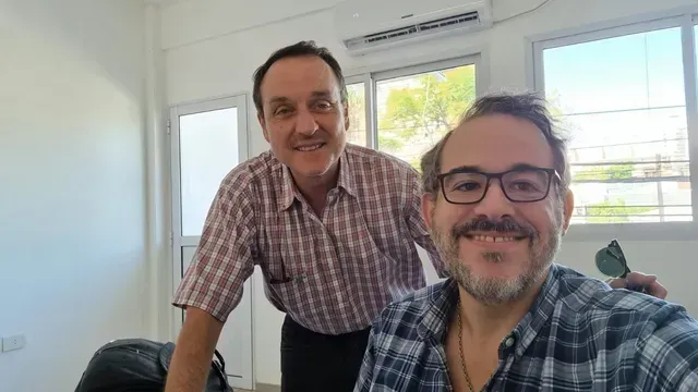Dos hombres sonrientes en un interior, uno con una camisa de cuadros, de pie junto a una ventana luminosa.