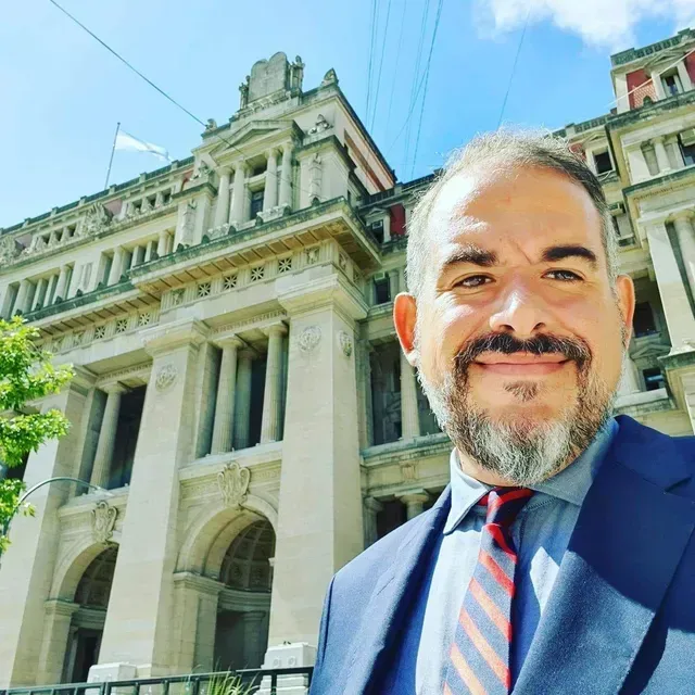 Un hombre con traje azul sonríe frente a un imponente edificio histórico bajo un cielo azul despejado