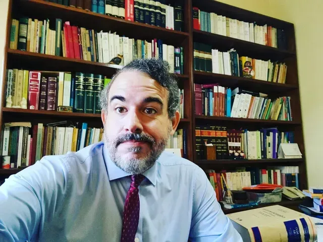 Persona sentada en una oficina, de frente a la cámara, con estanterías y libros de fondo