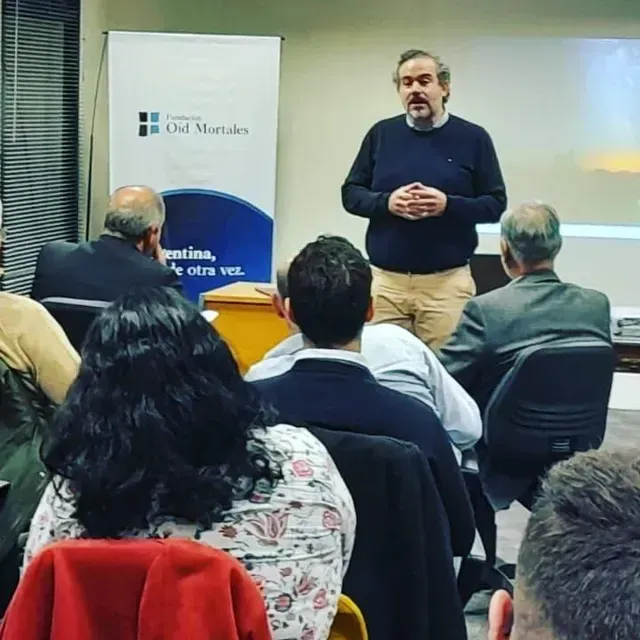 Un hombre habla ante un público sentado en un aula, con una pancarta y una pantalla de presentación detrás de él.