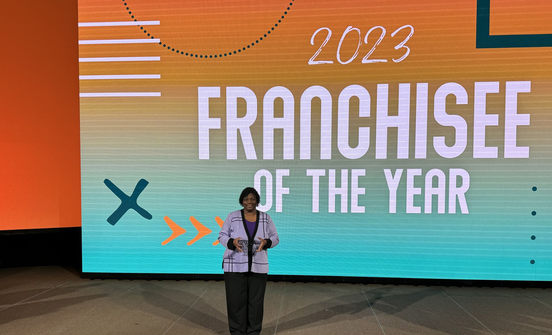 Person standing on stage in front of “2023 FRANCHISEE OF THE YEAR” screen with orange and teal graphics