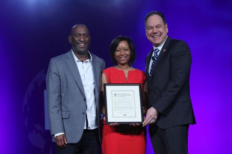 Three people onstage holding a framed award certificate against a purple-lit backdrop.