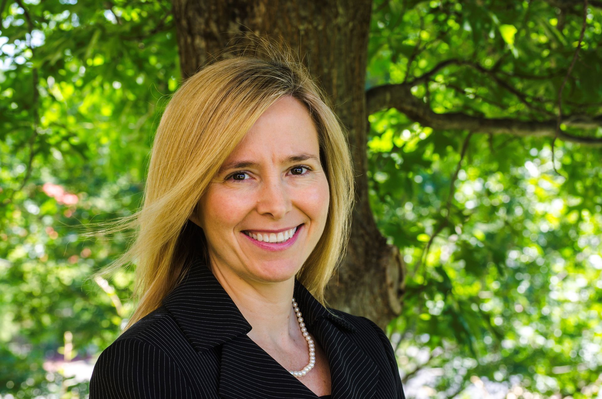 Smiling woman in a black top standing outdoors in front of a tree and green foliage