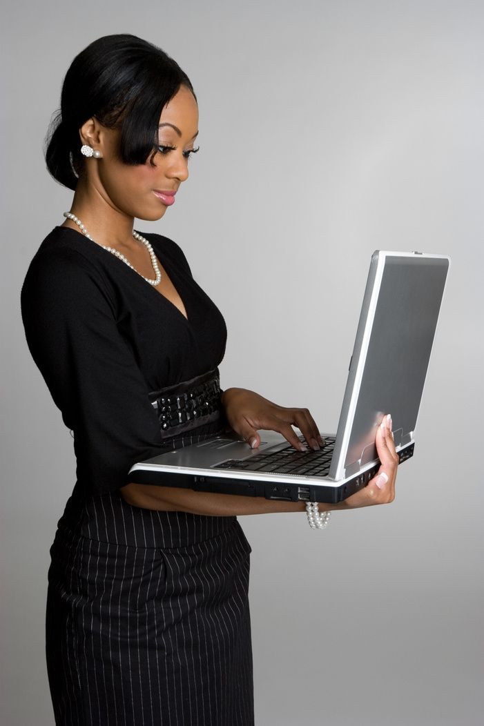 Woman in black attire holding and using a laptop; focused expression, neutral background.