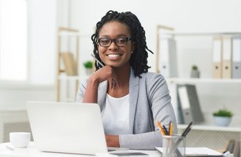 Woman wearing glasses smiles at the camera, sitting at a desk with a laptop.