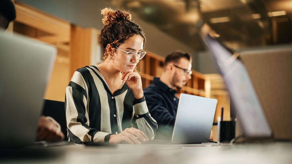 Woman in glasses, striped top, typing on laptop in an office, other people and laptops in the background.