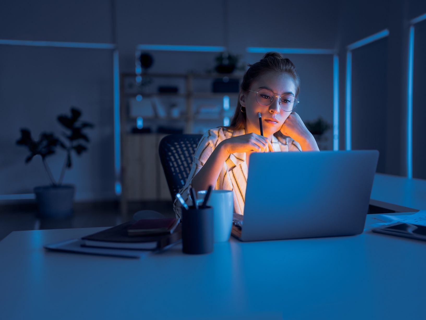 Woman working late at night on a laptop, in a dimly lit office. She holds a pen, looking intently at the screen.