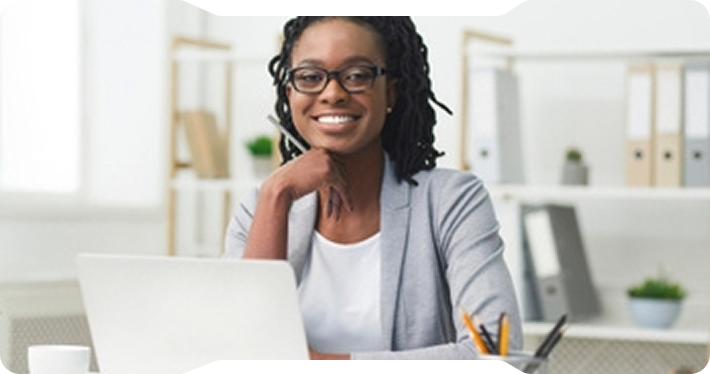 Woman smiling at laptop, leaning on hand. In office, wearing glasses and blazer.