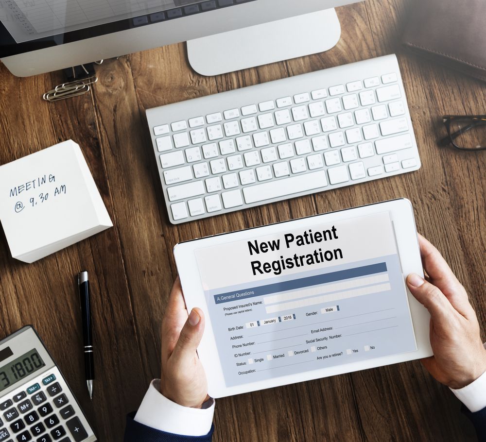 Hands filling out a New Patient Registration form at a desk with keyboard, calculator, and notebook