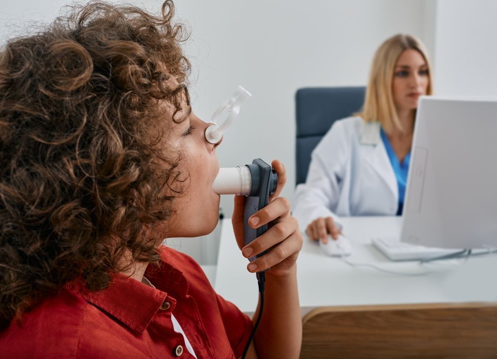Child using an inhaler while a doctor looks on at a desk in a clinic