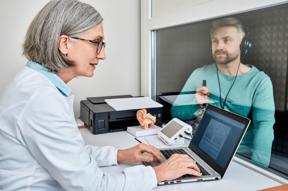 Doctor consulting with a man through a glass partition, using a laptop and office phone on a desk.