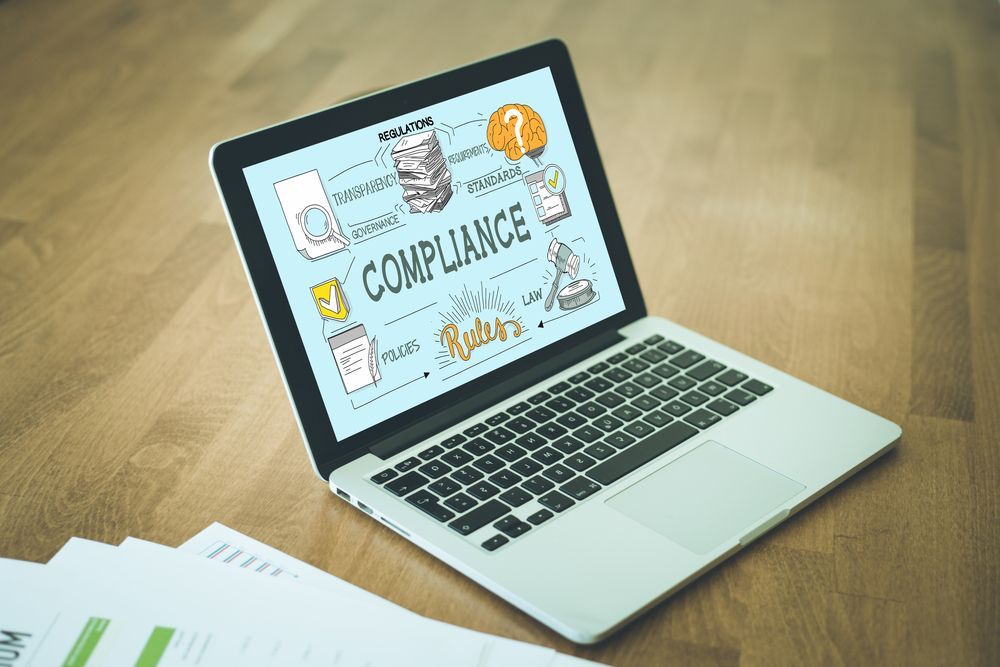 Laptop on desk displaying “COMPLIANCE” presentation with charts and icons, beside a paper report