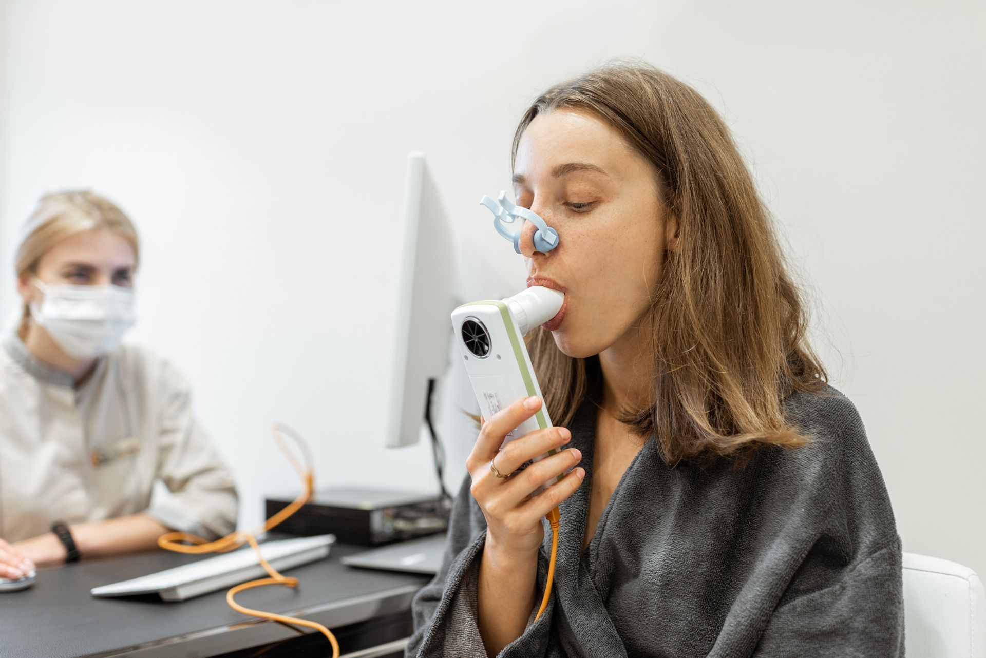 Person using a spirometer while another masked person sits nearby in a medical office