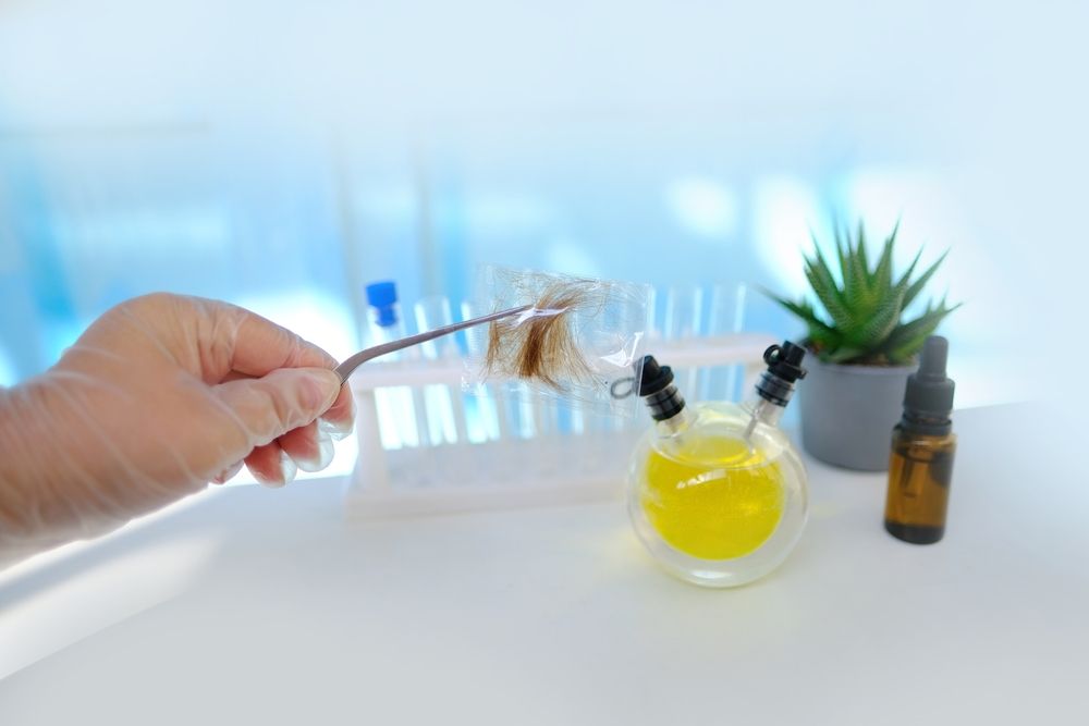 Hand holding a glass dropper above a yellow liquid next to a plant and amber bottle on a white table