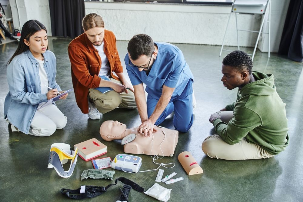 People kneeling around a CPR mannequin during first-aid training, with medical kits on the floor.