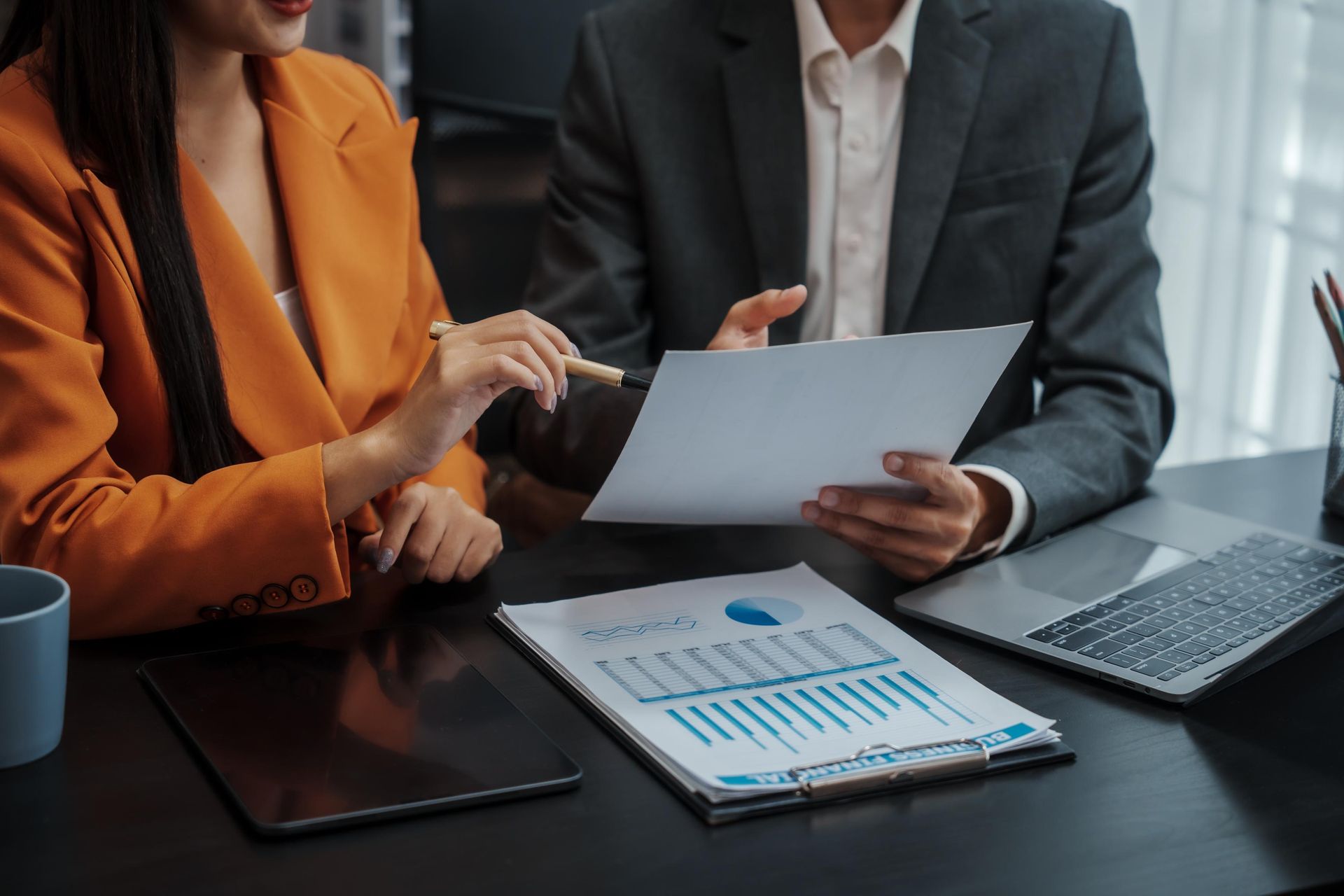 Two people in business attire review charts and a document at a desk.