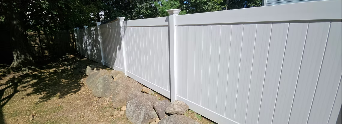 White vinyl privacy fence along a dirt path beside trees and a house, in bright sunlight