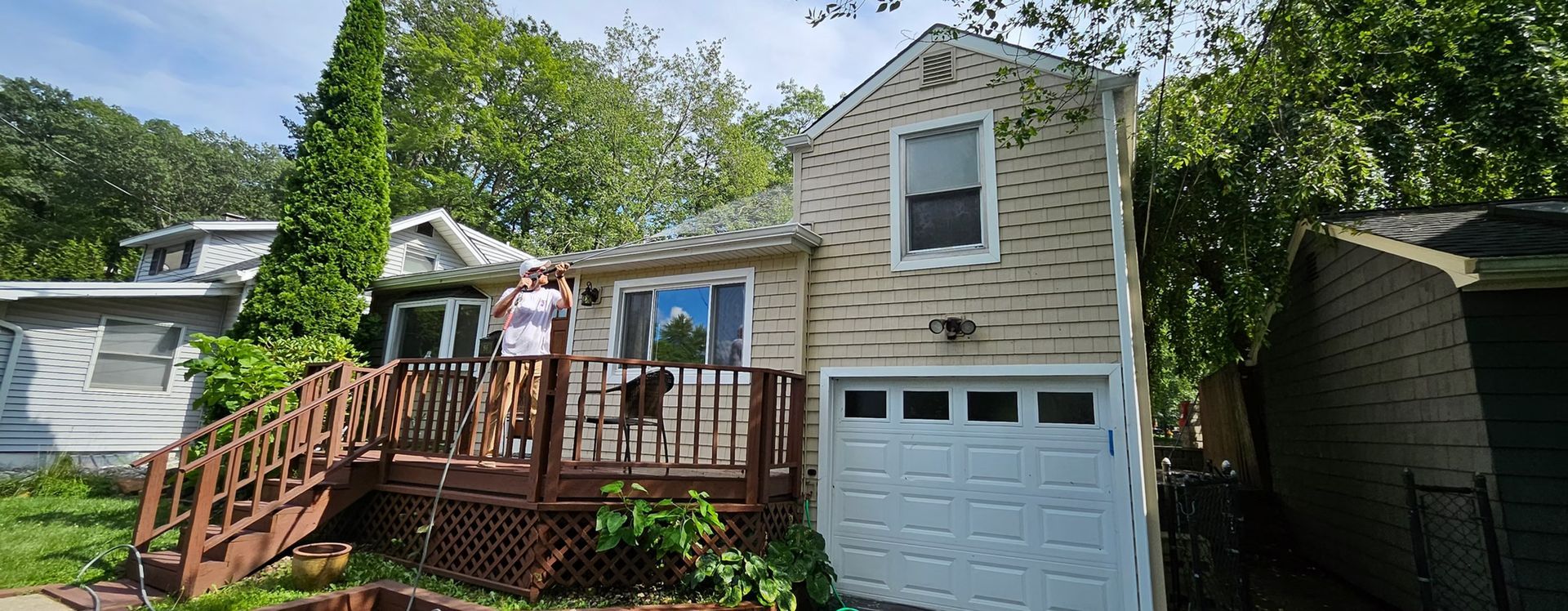 White house with wooden deck, stairs, and garage beneath trees on a sunny day