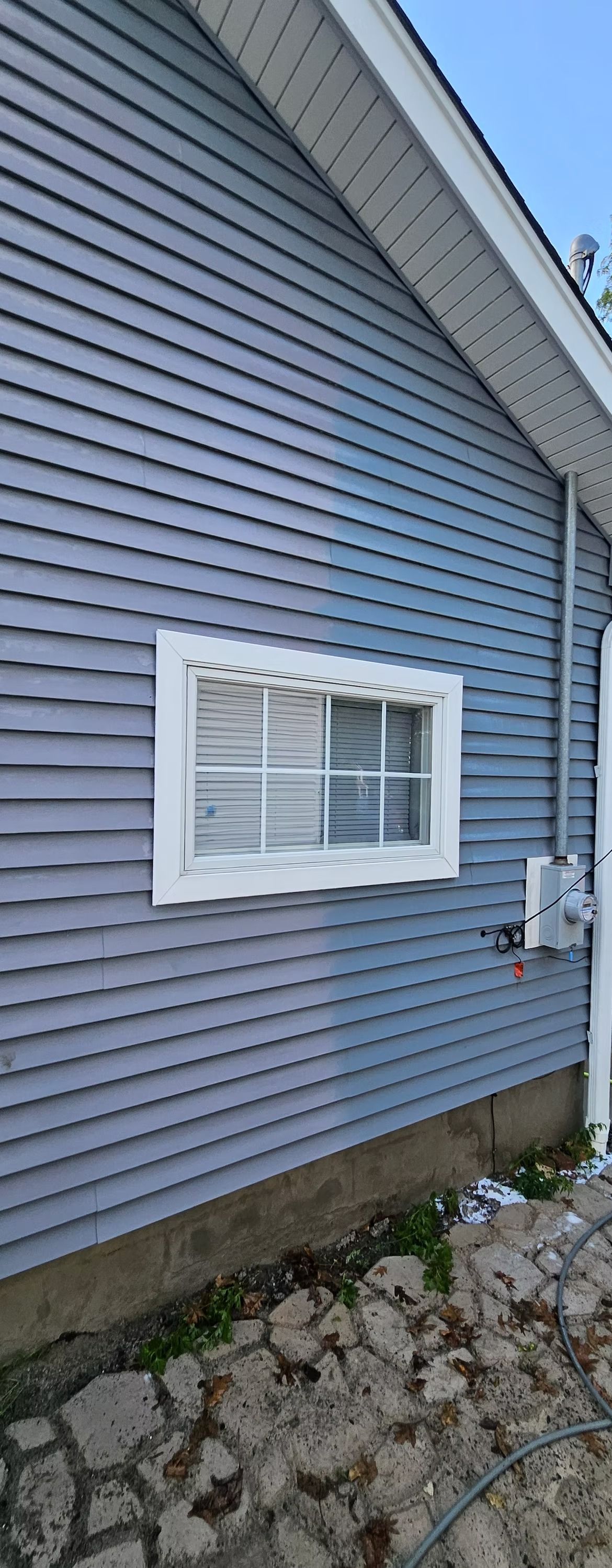 Blue house siding with a small white-framed window, utility meter, and rocky ground below.