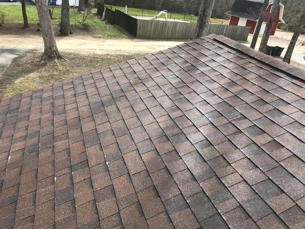 Brown shingled roof with trees and a fenced yard in the background