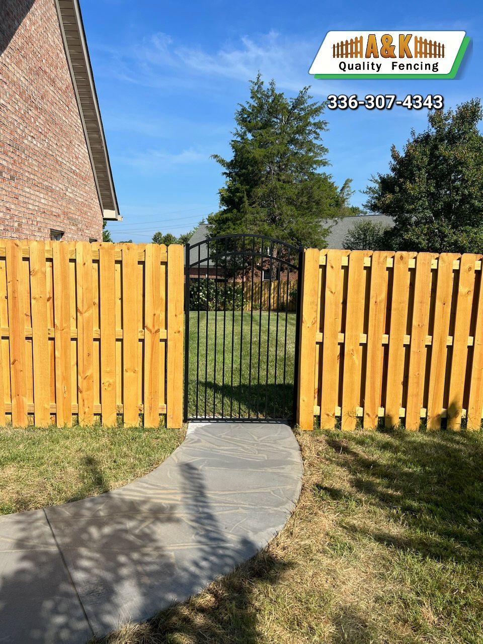 Wooden fence with black gate, concrete path, and brick building under a blue sky.