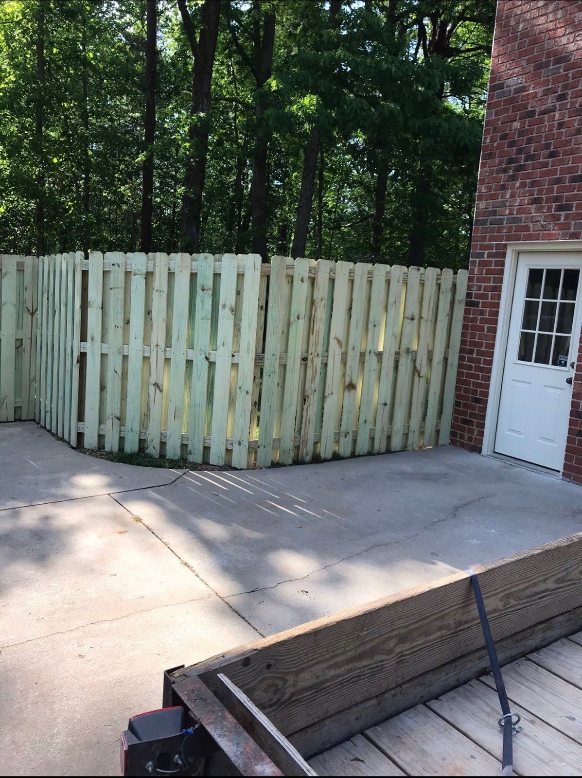 A new wooden fence surrounds a patio, beside a brick building and trees in the background.