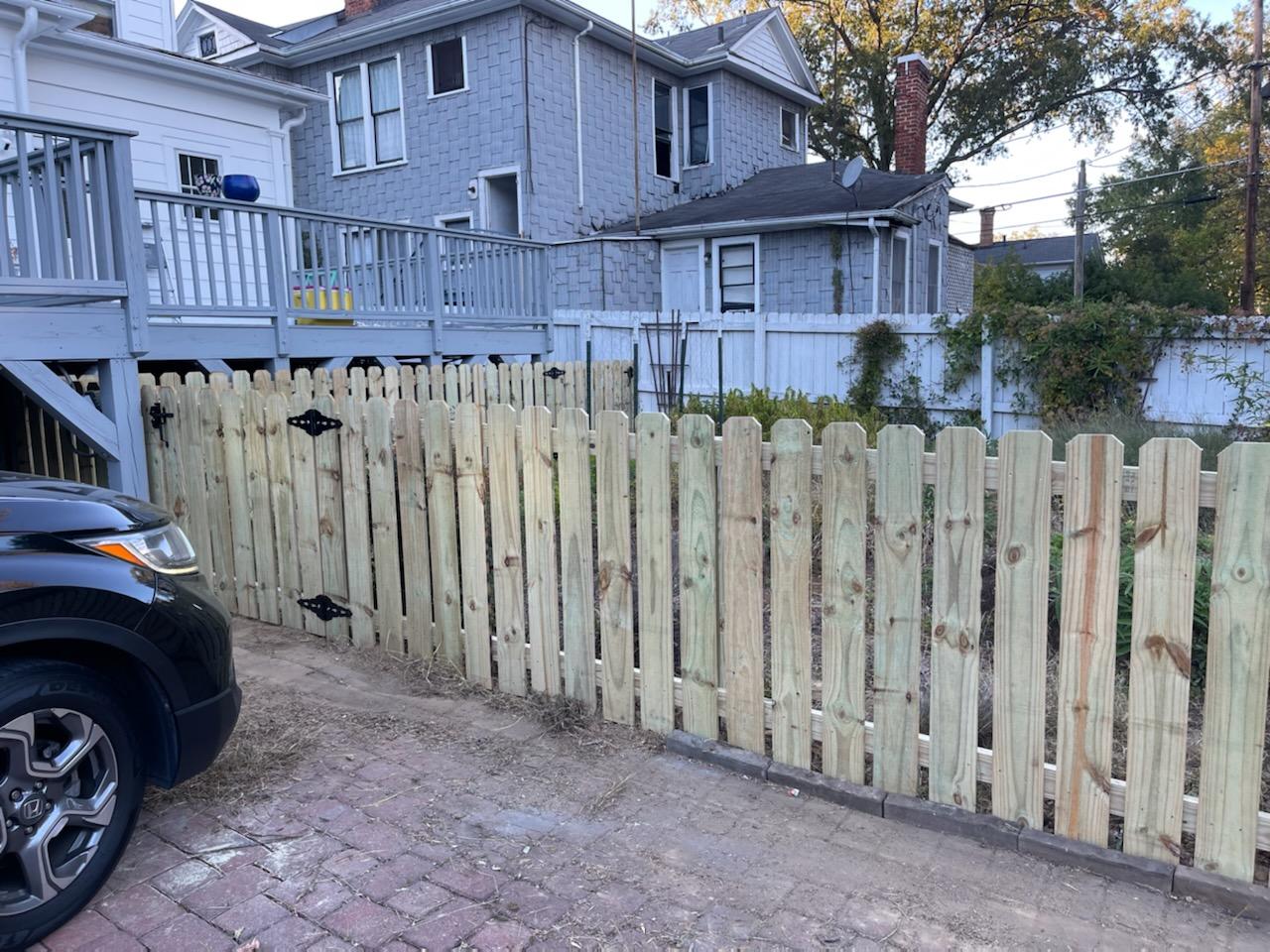 A newly built wooden picket fence in front of light-colored houses and a black car.