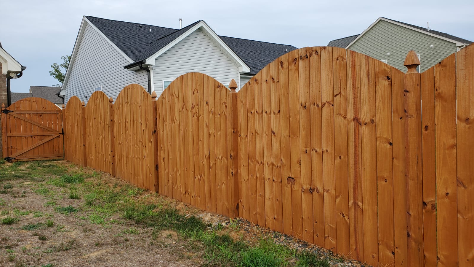 Wooden fence with arched tops, stained brown, in front of houses with gray roofs, and green grass.