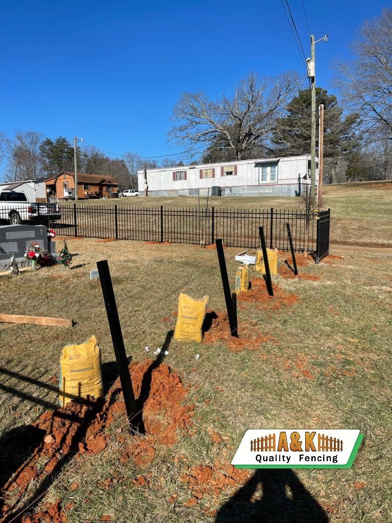 Fence posts set in holes with concrete bags, near a metal fence and mobile home on a sunny day.