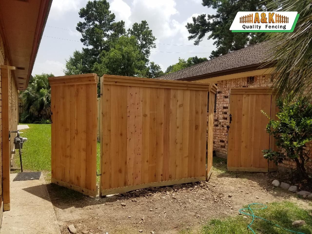 Wooden fence with two sections open, beside a brick house and vegetation.