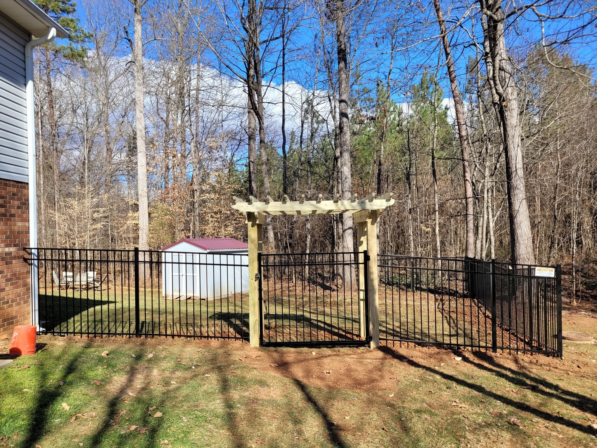 A black metal fence encloses a backyard with a pergola-covered gate, small shed, and trees.
