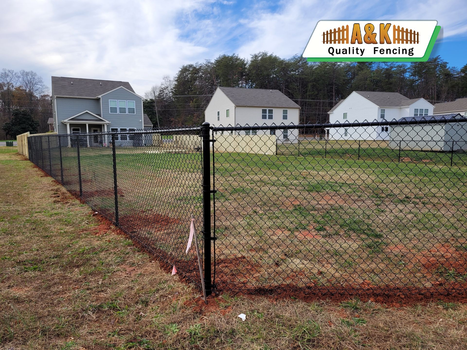 Black chain-link fence bordering a grassy area with houses in the background under a blue sky.