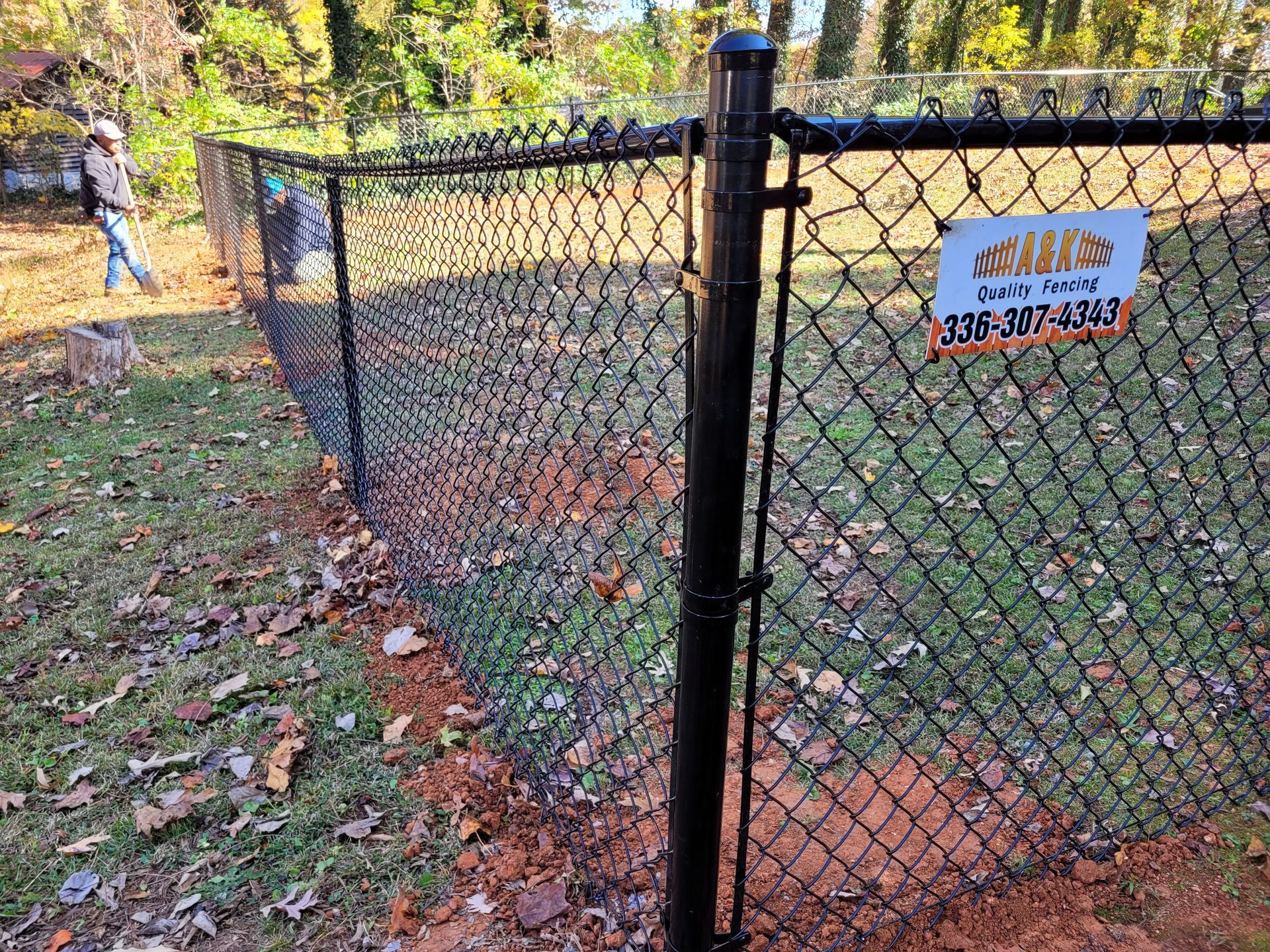 Black chain-link fence in grassy yard with fallen leaves. A person walks near the fence.