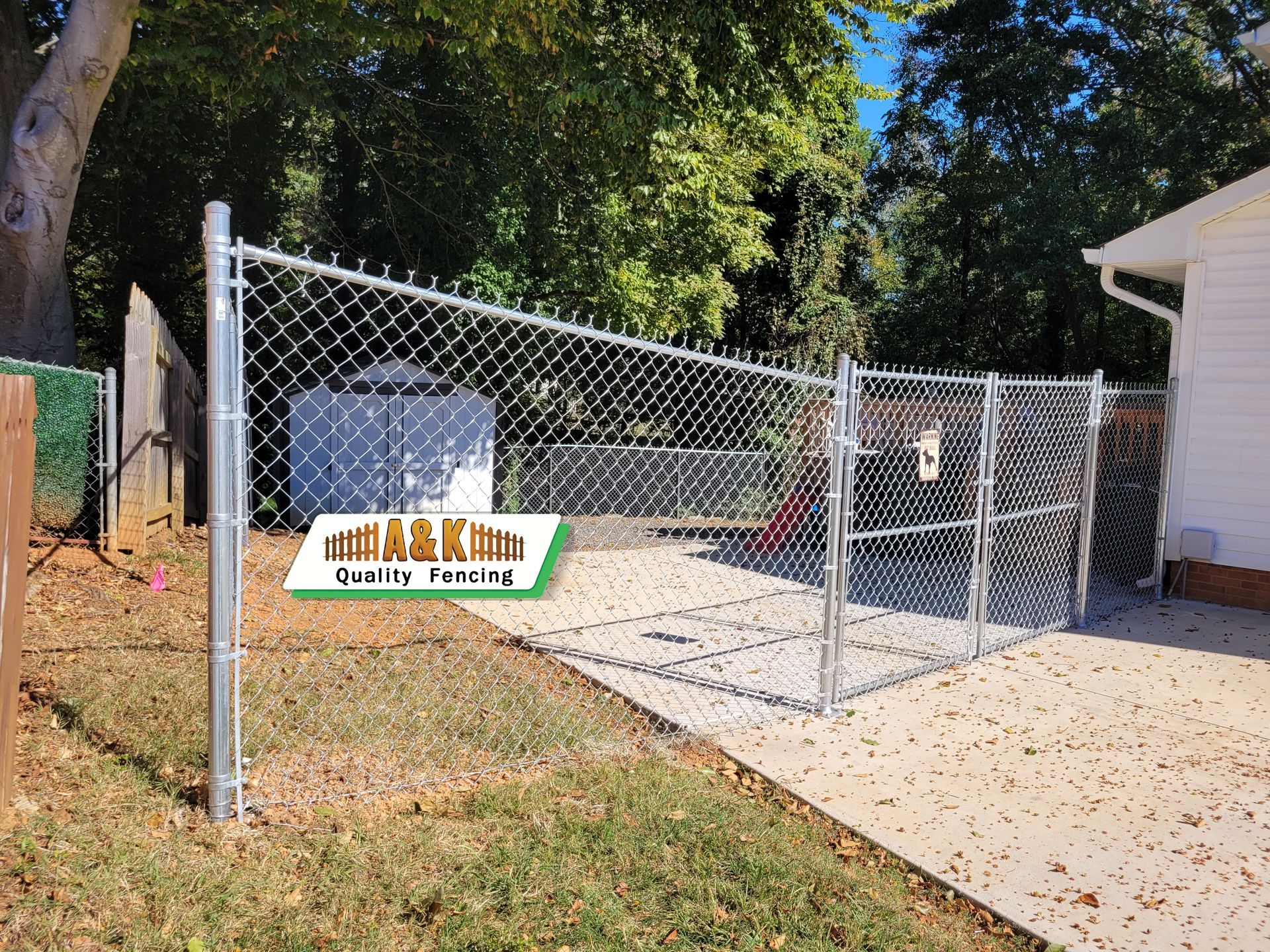 Chain-link fence encloses a concrete area next to a house with a green lawn. 